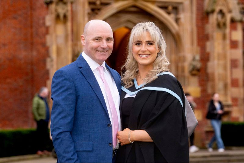 Image of Queen's University Belfast's Head Porter, Martin Mooney, pictured alongside his daughter in her graduation gown in front of the red brick Lanyon building